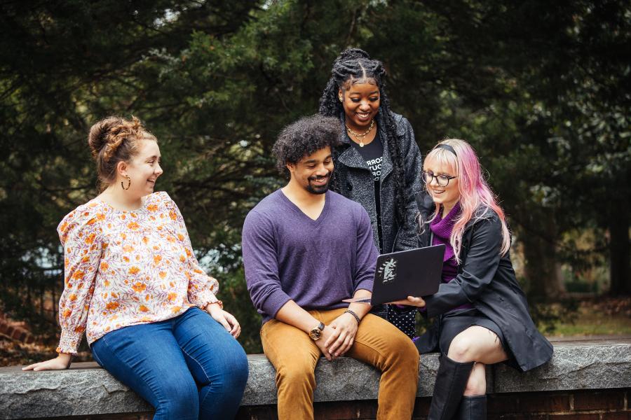 group of students sit on a ledge in fall clothing while looking at a laptop screen