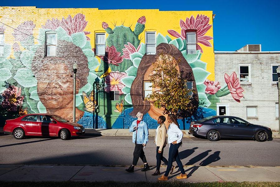 group of three students walking down sidewalk in front of bright mural of two faces